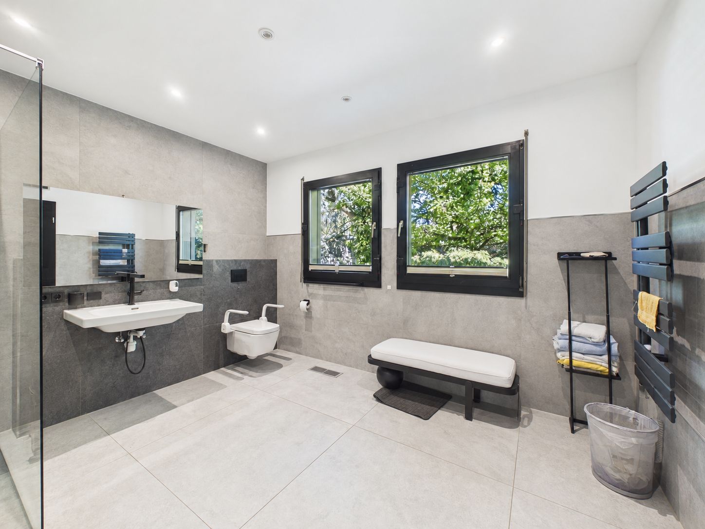 Modernes, geräumiges Badezimmer mit Waschbecken, WC, Bank, zwei Fenstern und Regal mit Handtüchern.