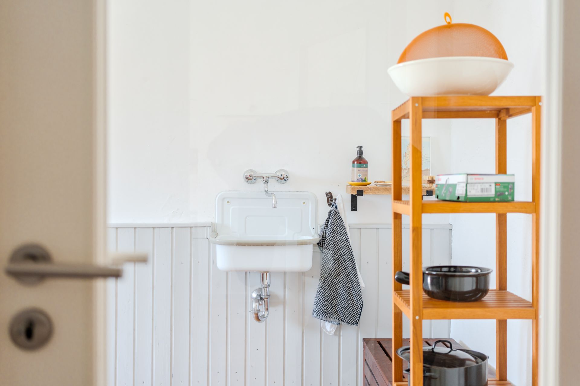 Kleines Waschbecken mit Handtuch, Seife und Holzregal mit Schalen und Töpfen in einem hellen Raum.
