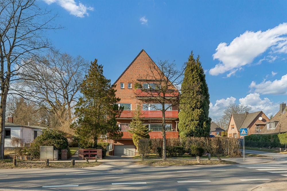 Dreistöckiges Wohnhaus mit rotem Giebel und Balkon, Bäume und Straße im Vordergrund.