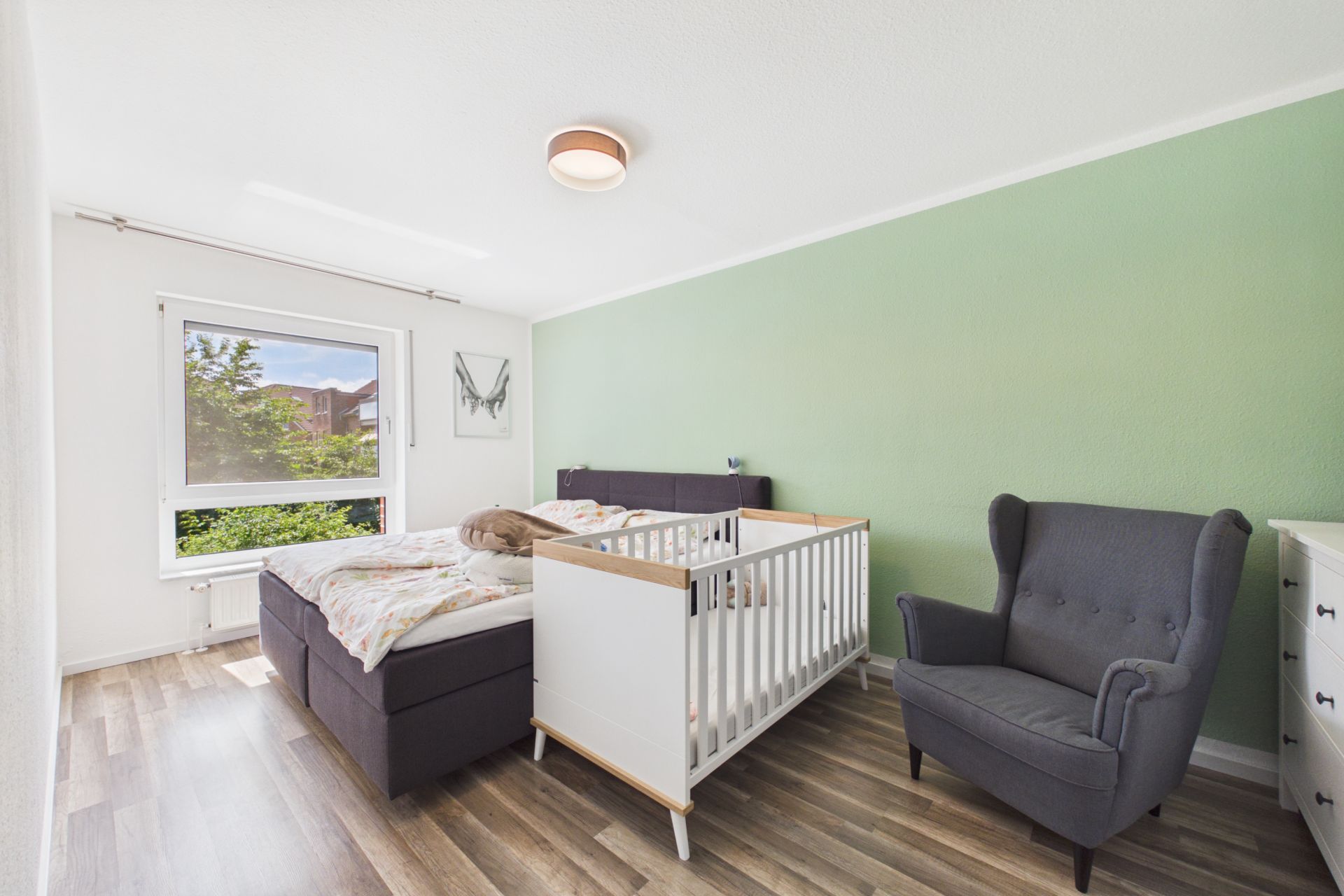 Helles Schlafzimmer mit Doppelbett, Babybett, Sessel und grün gestrichener Wand.