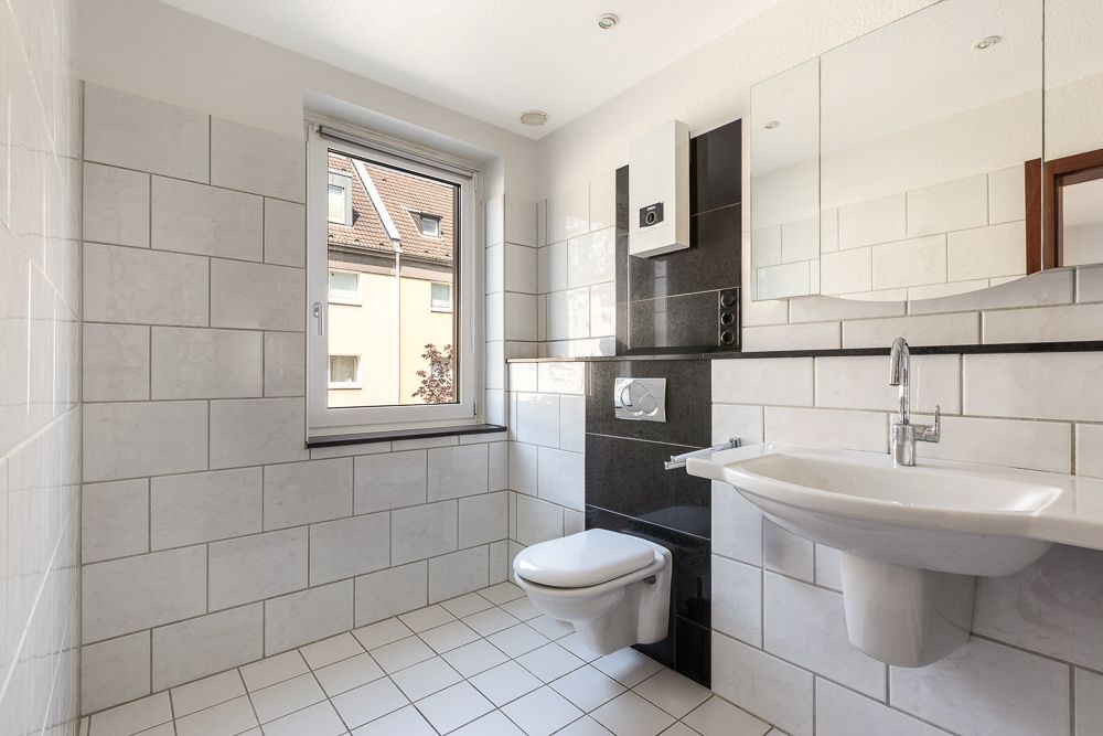 Helles Badezimmer mit Wand-WC, Waschbecken und Fenster, weiße Fliesen an Wand und Boden.