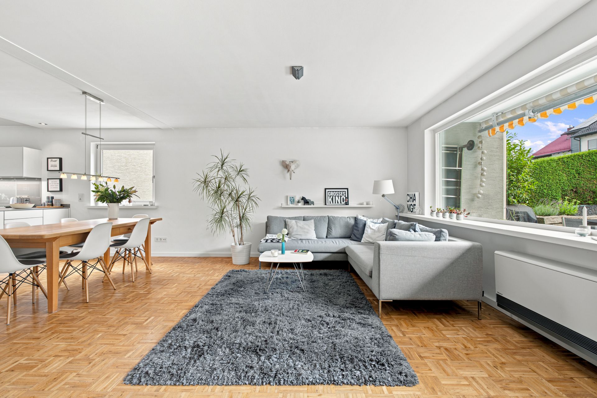 Modernes Wohnzimmer mit grauem Sofa, Teppich, Esstisch und großem Fenster mit Blick nach draußen.