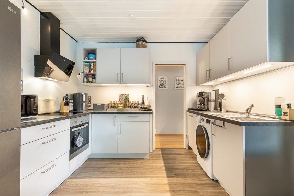 Moderne Einbauküche mit weißen Schränken, Elektrogeräten und Waschmaschine auf Holzfußboden.