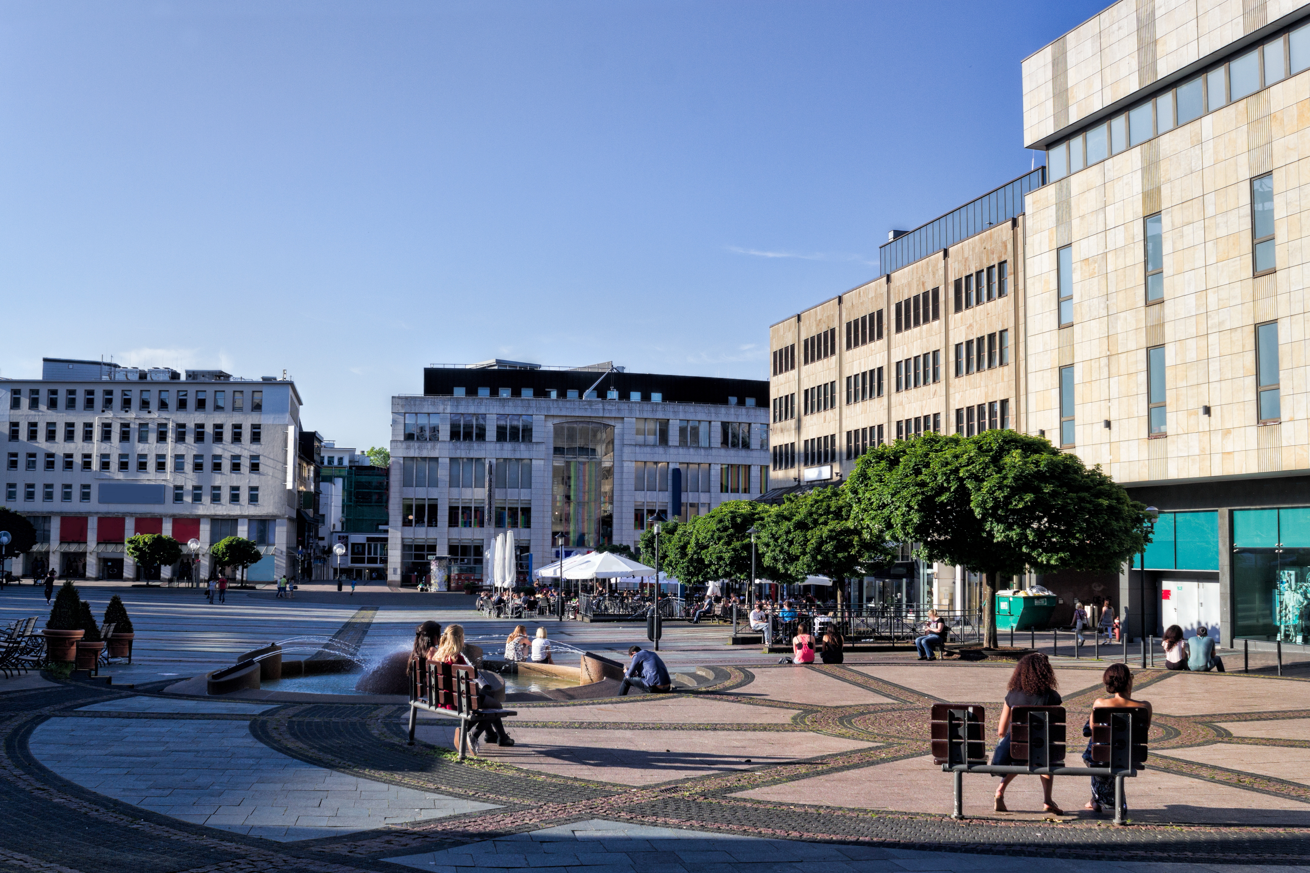 Stadtplatz mit Bänken, Bäumen und Menschen bei sonnigem Wetter.