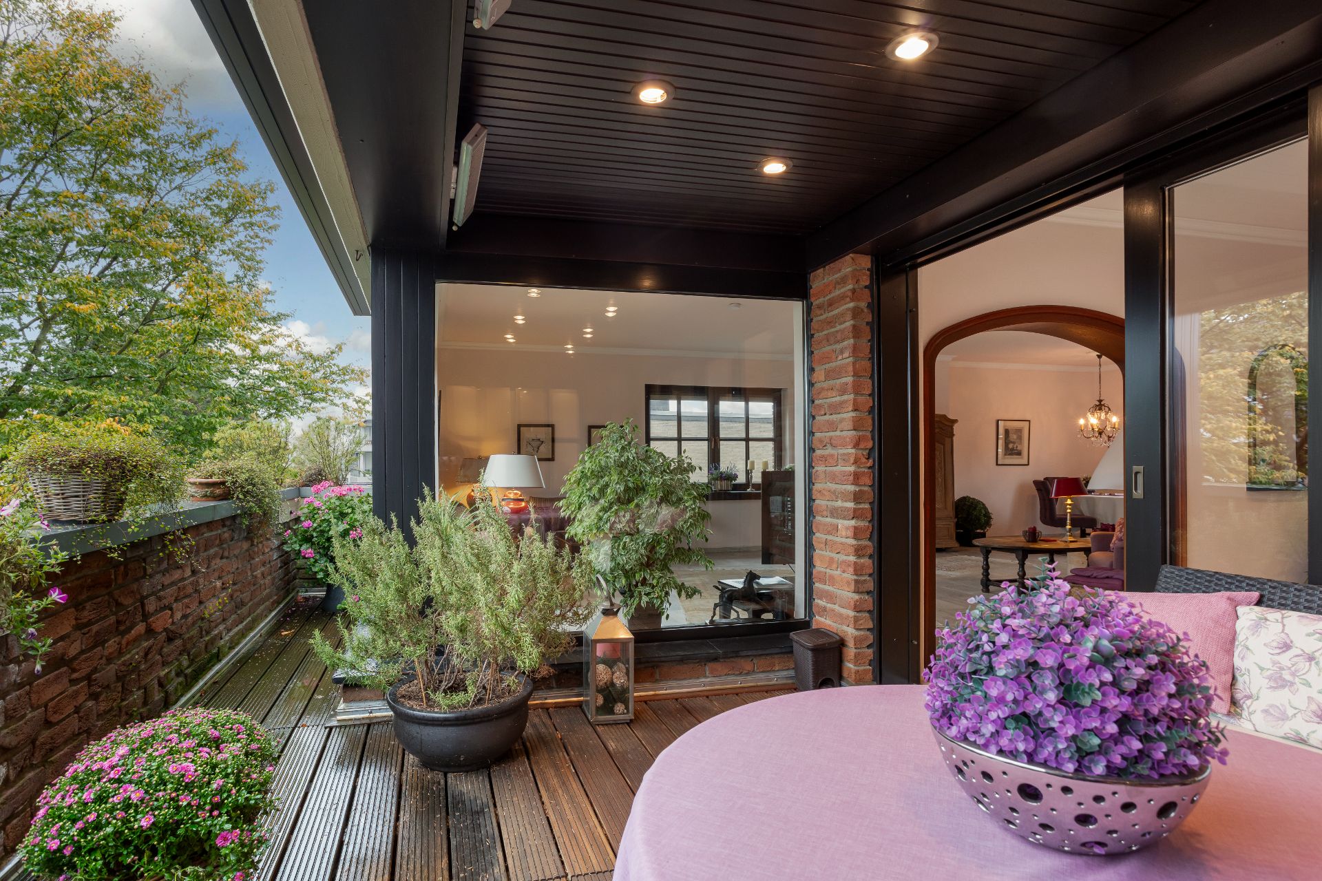 Überdachte Terrasse mit Holzboden, Pflanzen und Blick in ein Wohnzimmer mit großen Fenstern.