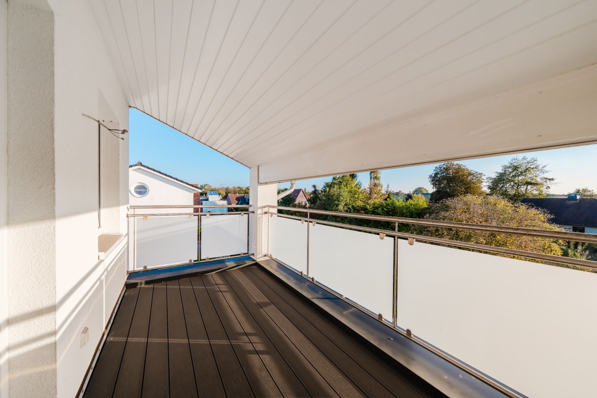 Überdachter Balkon mit Holzboden und Glasgeländer, Blick auf Bäume und Nachbarhäuser.