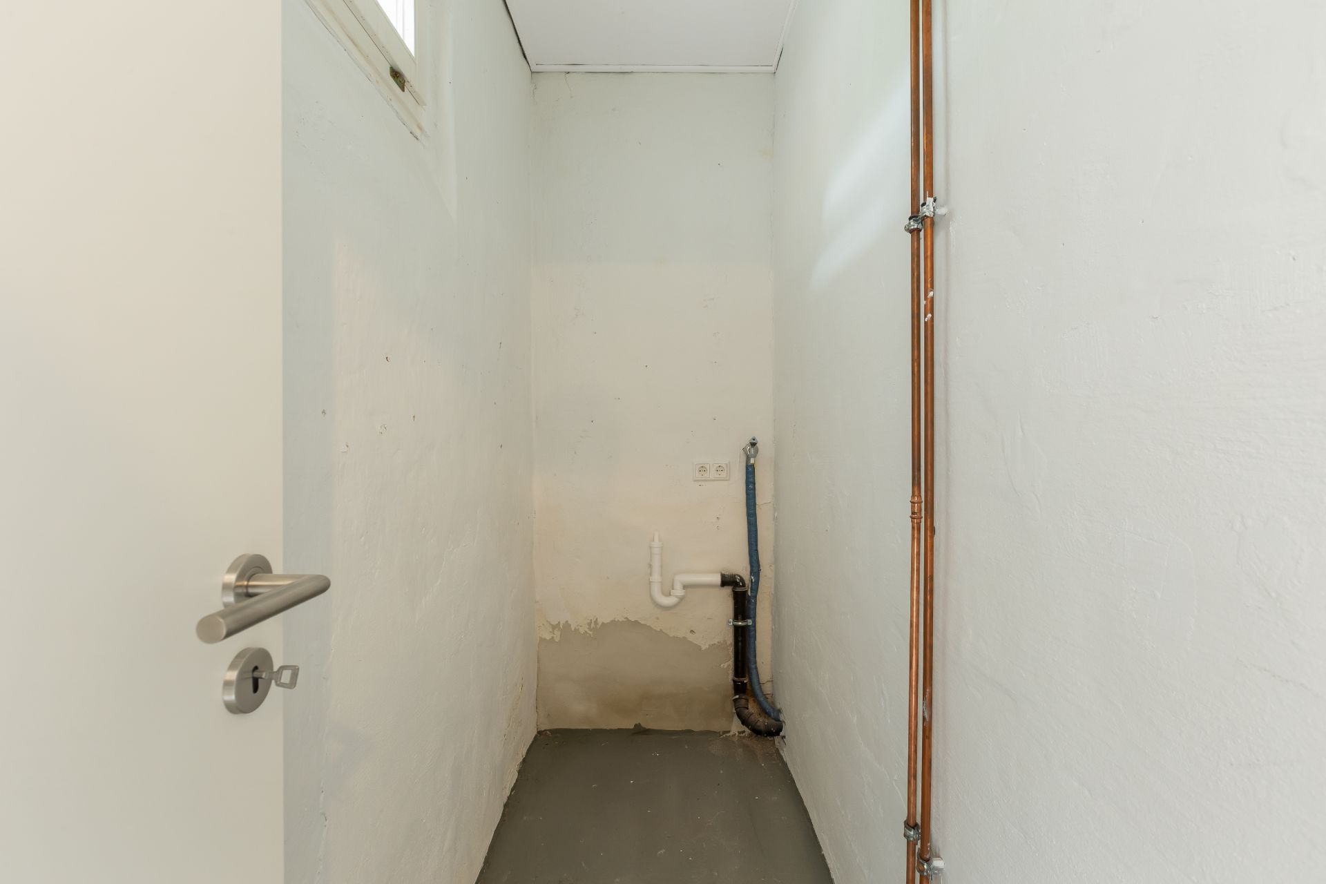Kleiner leerer Raum mit weißen Wänden, Rohrleitungen und Anschlüssen an der Rückwand.