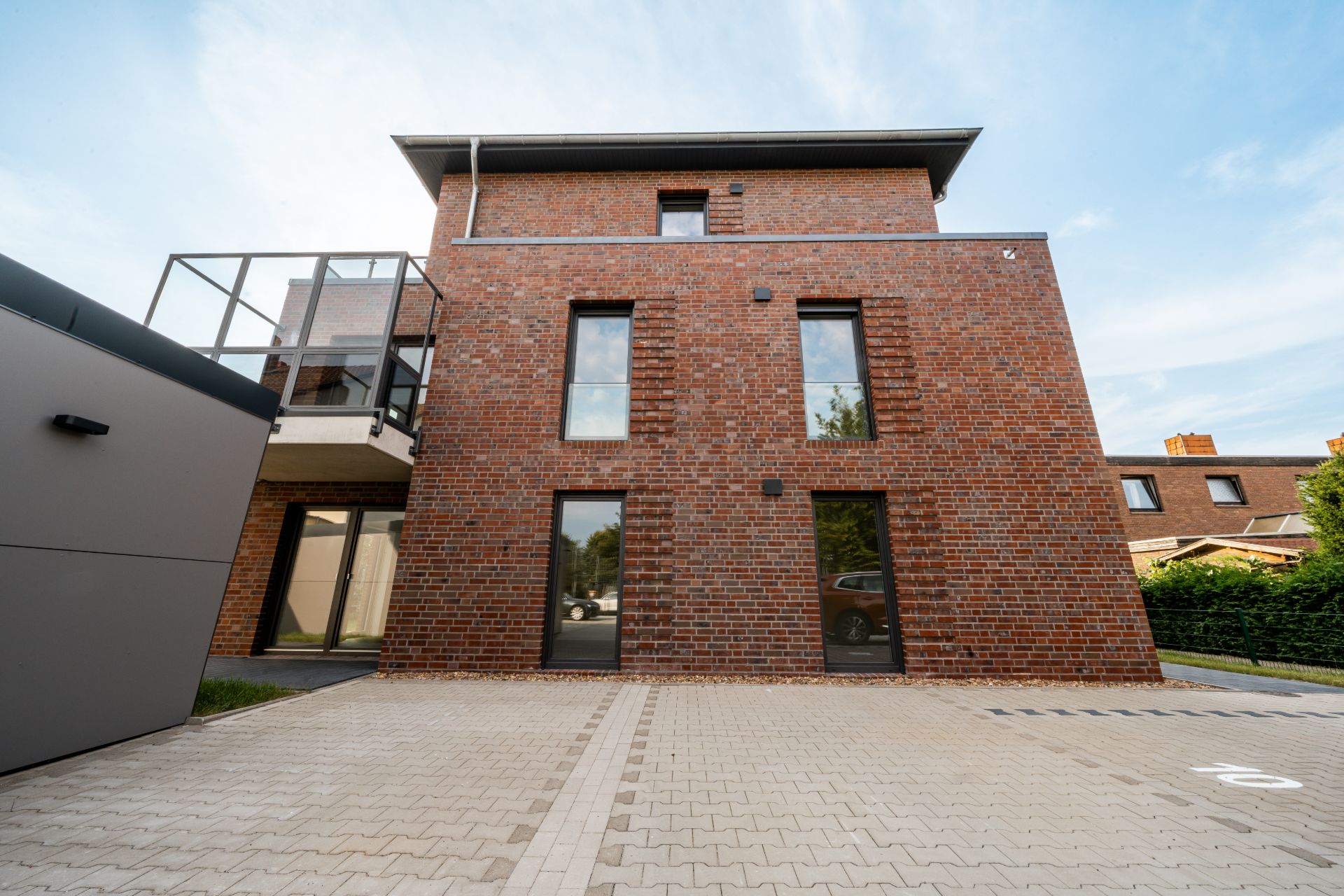 Moderne Wohnhausfassade aus rotem Klinker mit großen Fenstern und gepflastertem Parkplatz davor.