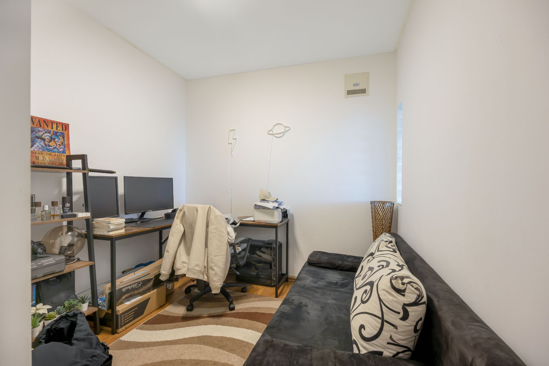 Kleines Arbeitszimmer mit Schreibtisch, zwei Monitoren, Regal und einem Sofa mit Kissen.
