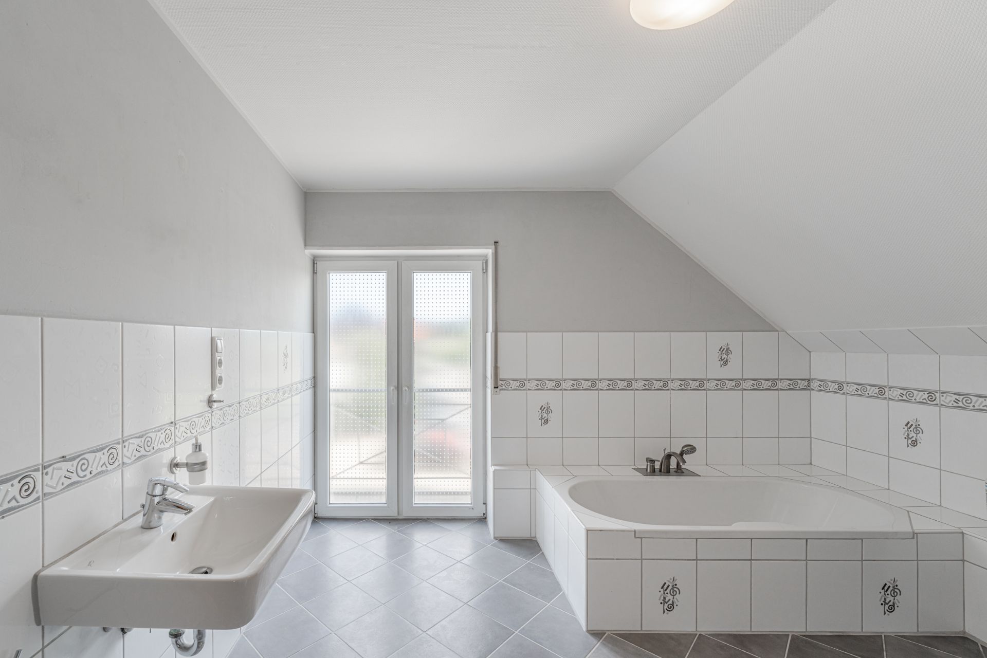 Helles Badezimmer mit Eckbadewanne, Waschbecken und Fliesen, Fenster im Hintergrund.