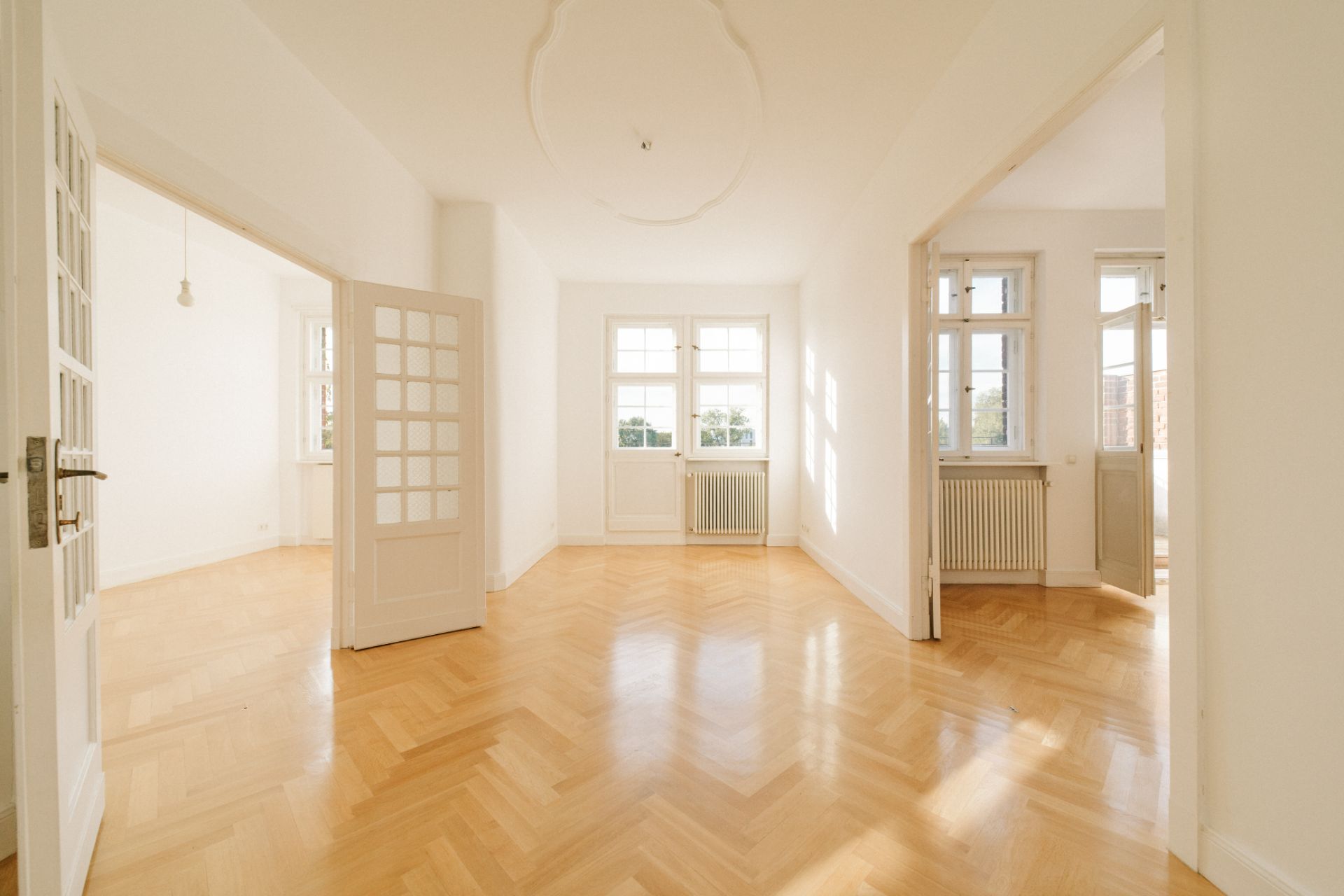Heller Raum mit Parkettboden, weißen Wänden und großen Fenstern, Türen führen in weitere Zimmer.