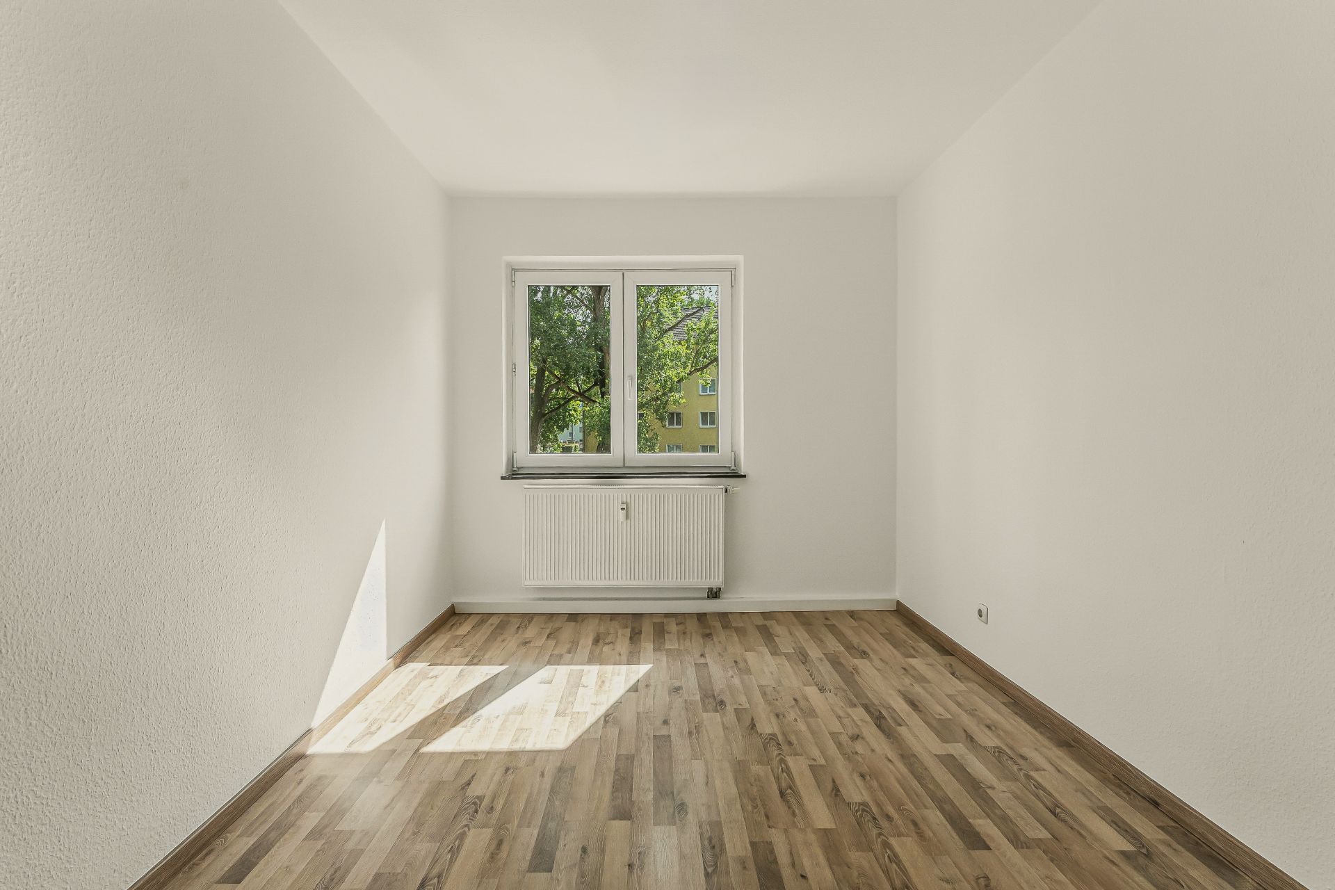 Leerer Raum mit Holzfußboden, weiß gestrichenen Wänden und einem Fenster mit Blick auf Bäume.
