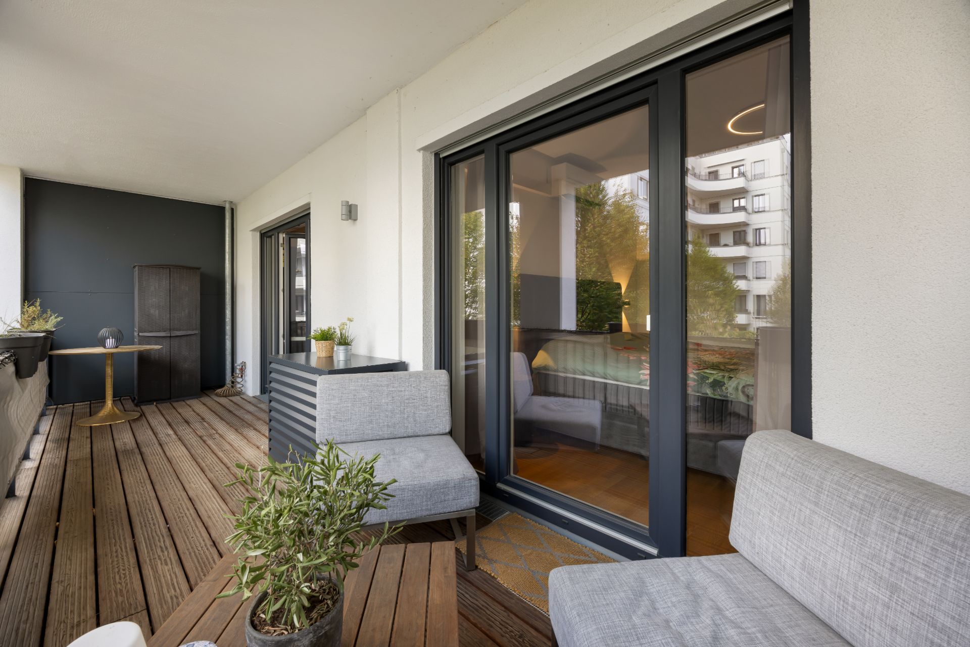 Überdachter Balkon mit Holzboden, Sitzmöbeln und Pflanzen vor einer großen Glas-Schiebetür.