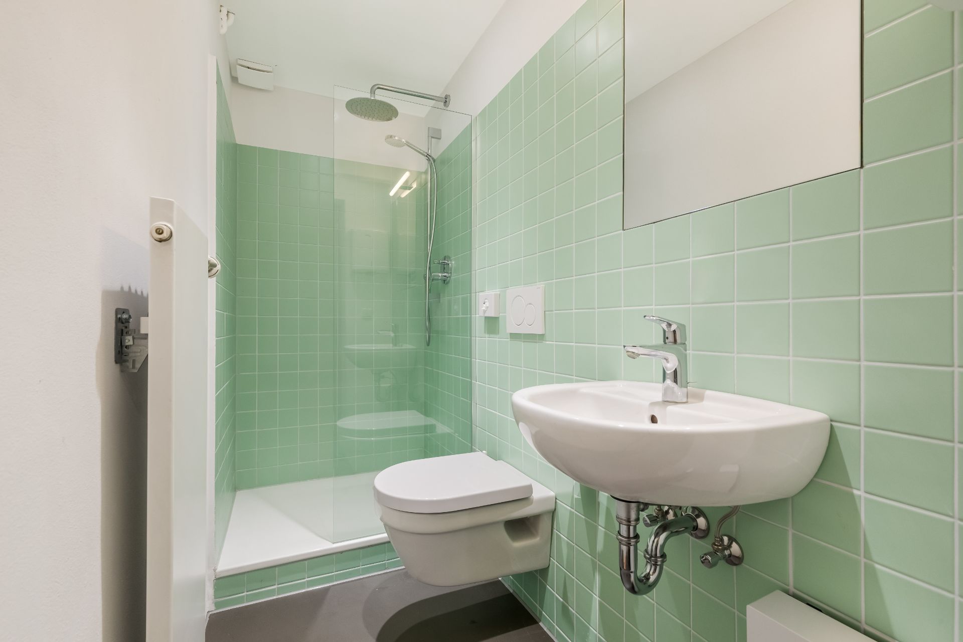 Modernes Bad mit grünen Fliesen, Wandtoilette, Waschbecken und offener Dusche.