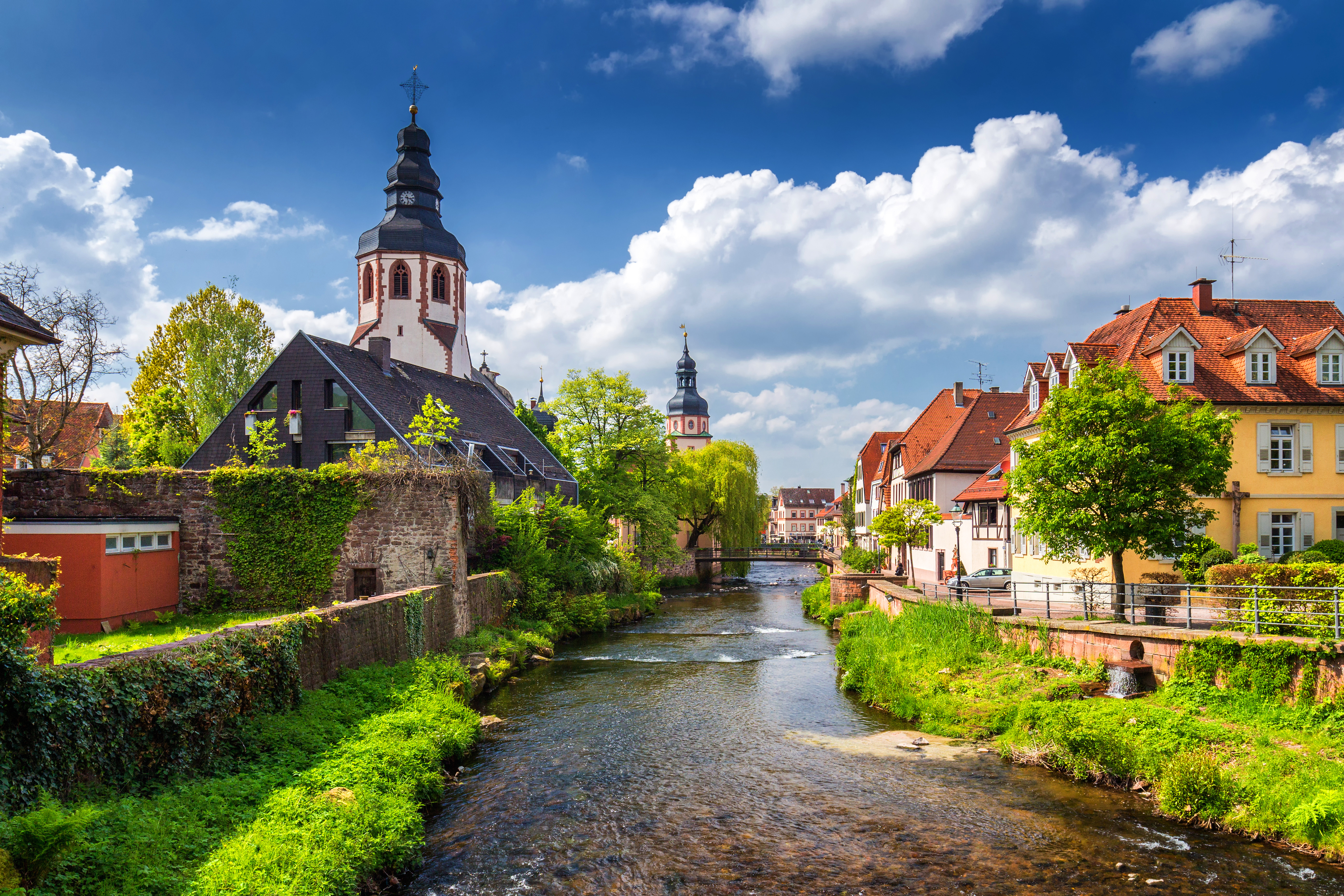 Fluss mit historischen Wohnhäusern und Kirche im Hintergrund unter blauem Himmel.