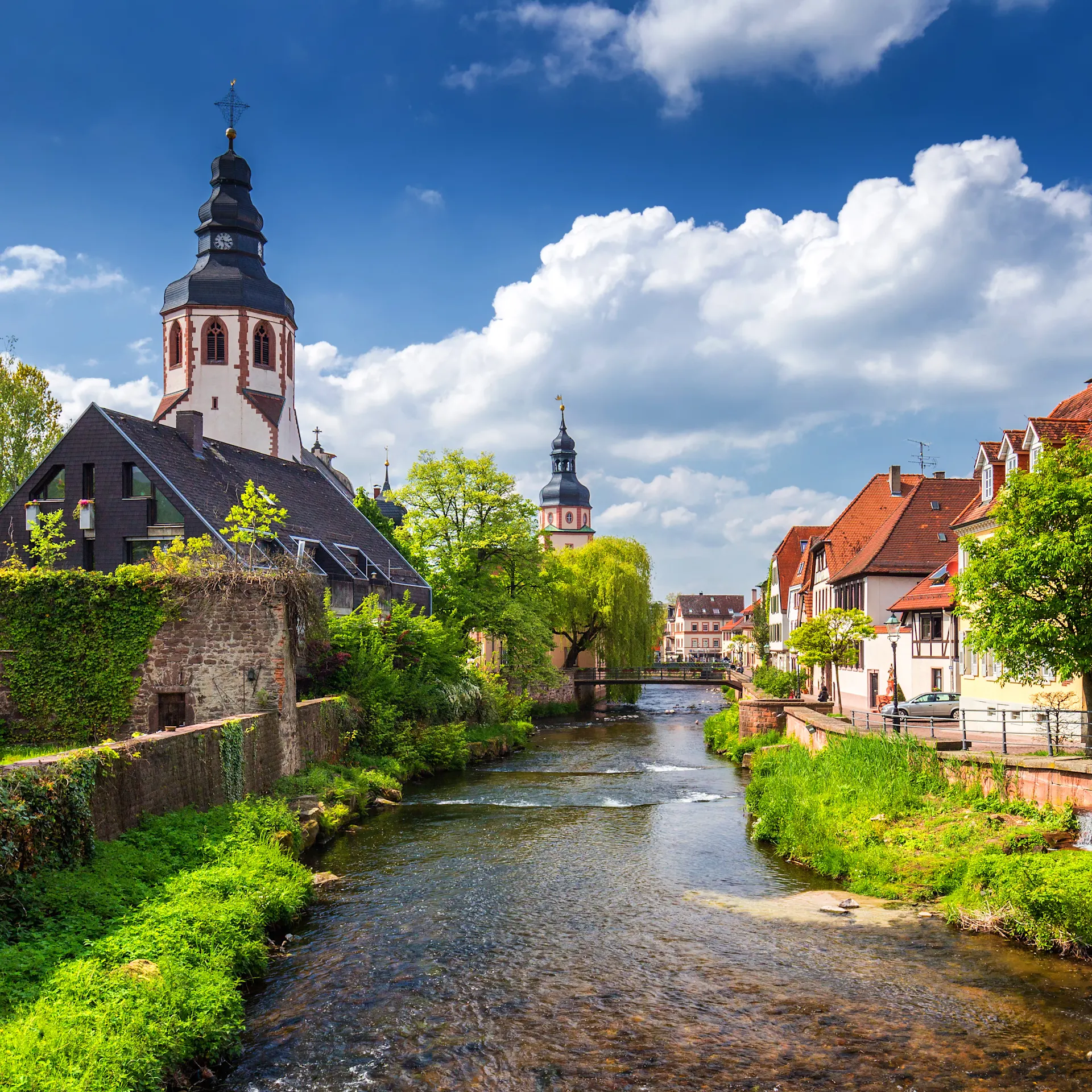 Fluss mit historischen Wohnhäusern und Kirche im Hintergrund unter blauem Himmel.