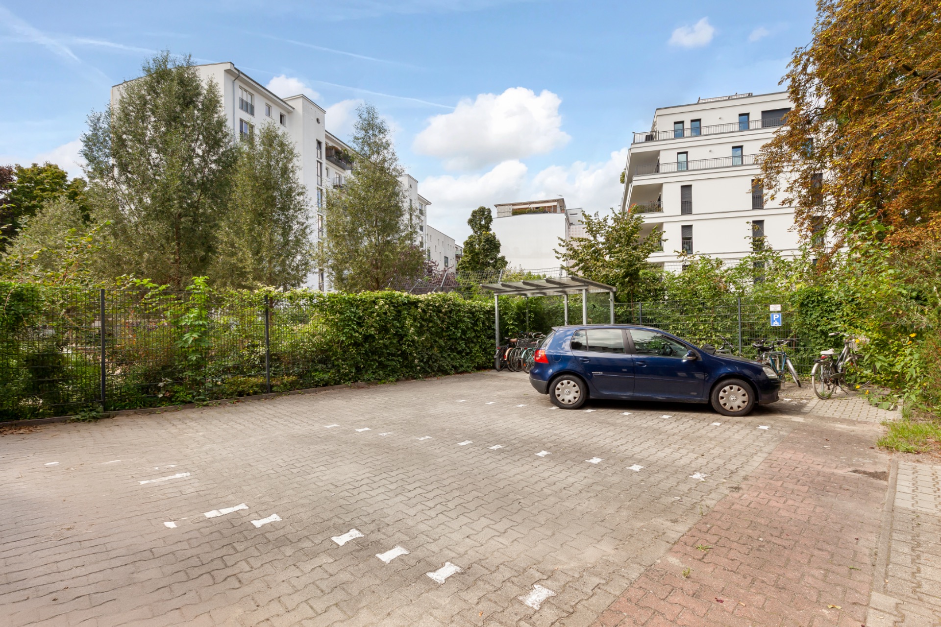 Parkplatz mit blauem Auto, daneben Fahrradständer und mehrstöckige Wohnhäuser im Hintergrund.