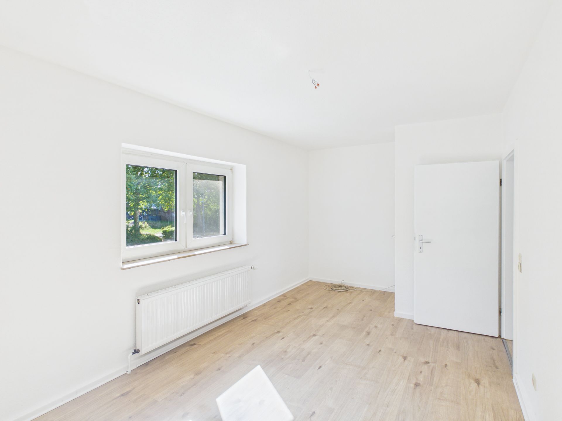 Heller, leerer Raum mit Holzfußboden, weißer Wand, Fenster und offener Tür.