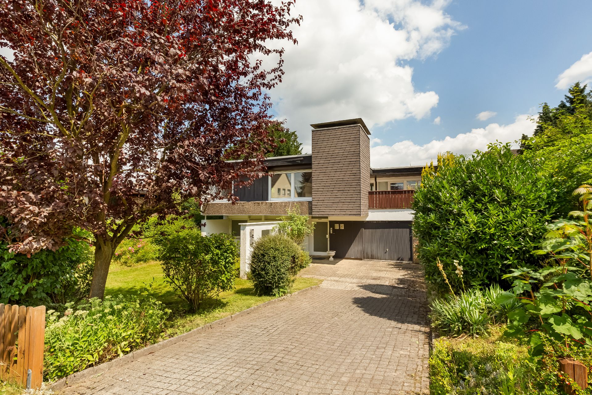 Einfamilienhaus mit gepflasterter Einfahrt, Garten und großem Baum im Vordergrund.