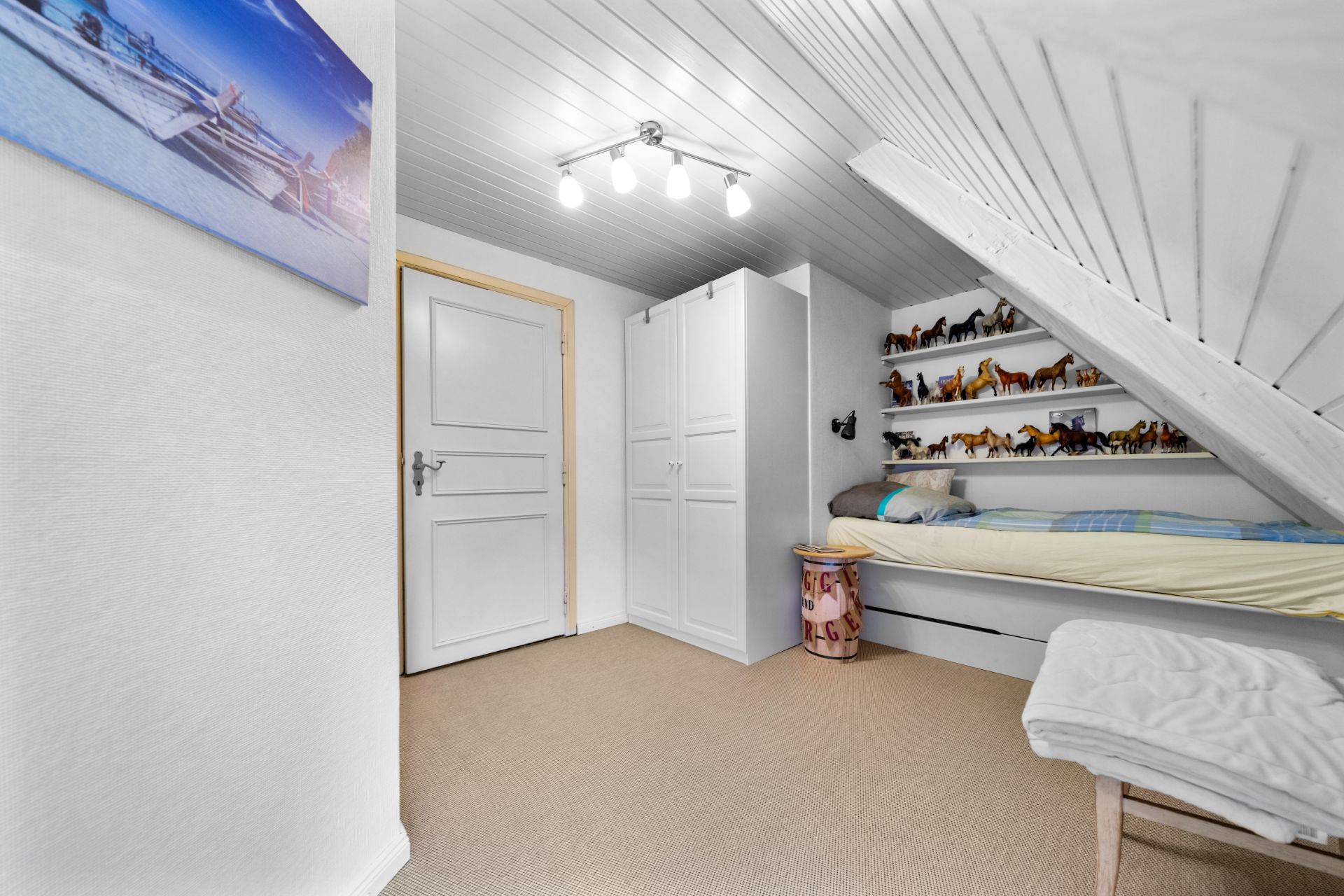 Kinderzimmer mit Einzelbett, Schrank, weißen Wänden und Regalen mit Spielzeugpferden unter einer Dachschräge.