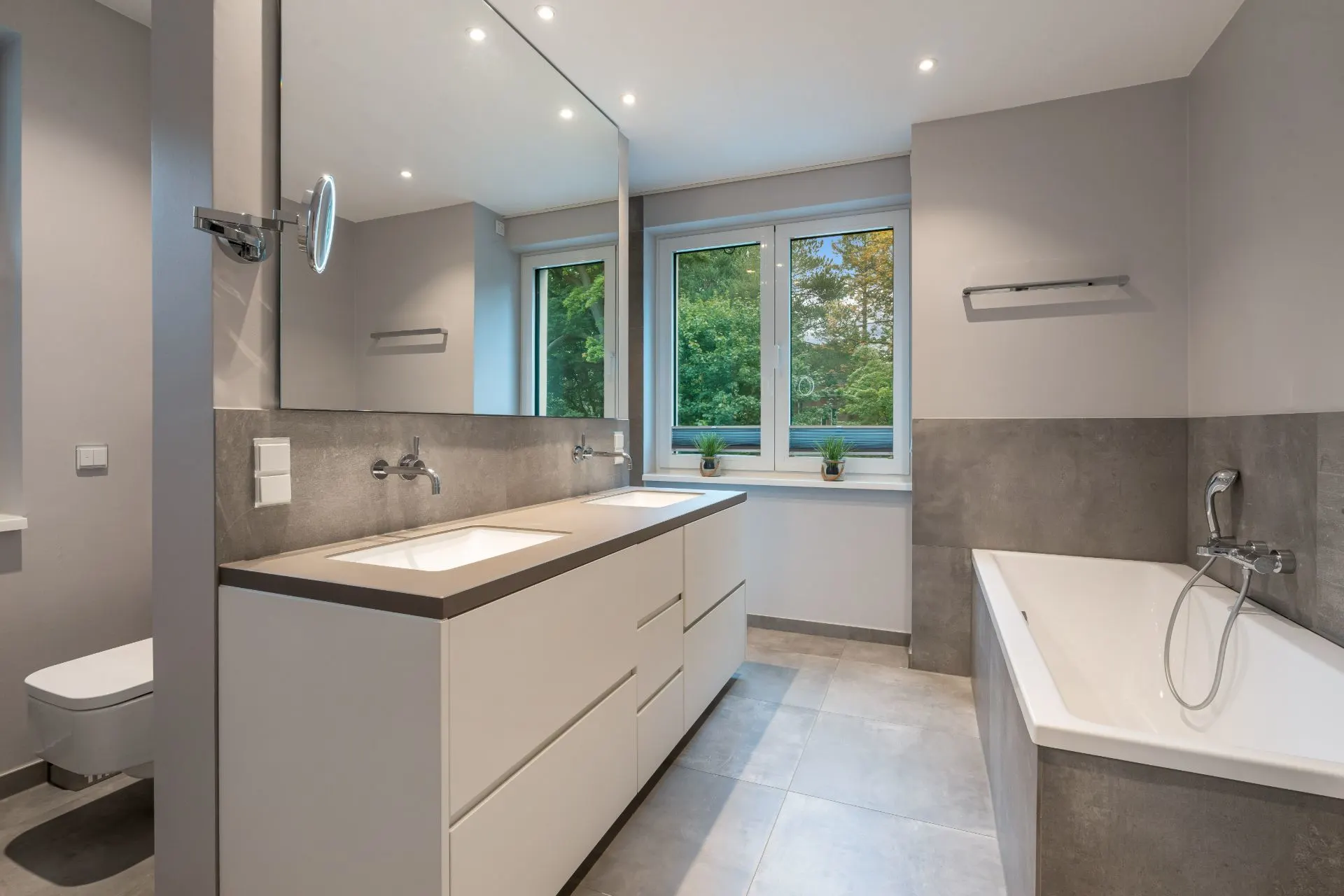 Modernes Badezimmer mit Doppelwaschbecken, großer Spiegel, Badewanne und Fenster mit Blick ins Grüne.