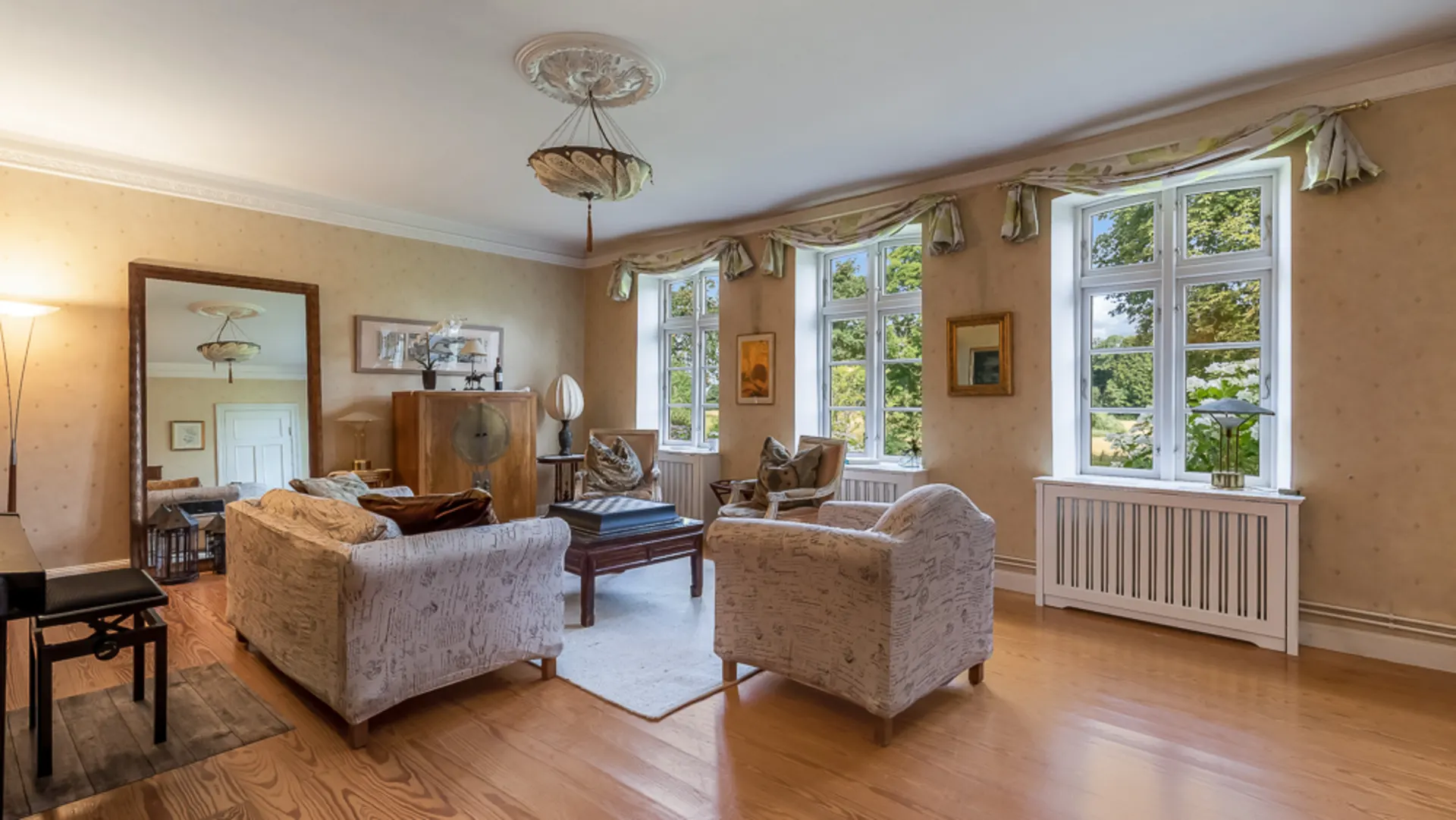 Helles Wohnzimmer mit Sesseln, Sofa, großem Spiegel und Fenstern mit Blick ins Grüne.