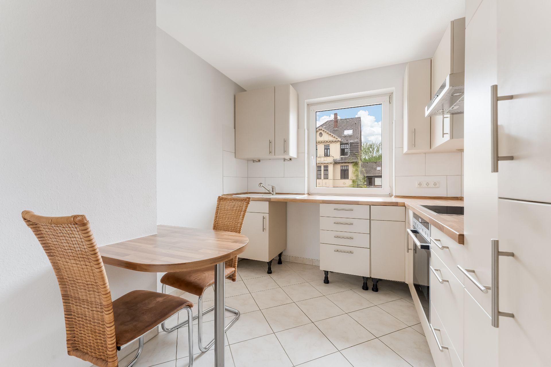 Helle Küche mit weißen Schränken, Holztisch und zwei Stühlen, Fenster mit Blick nach draußen.