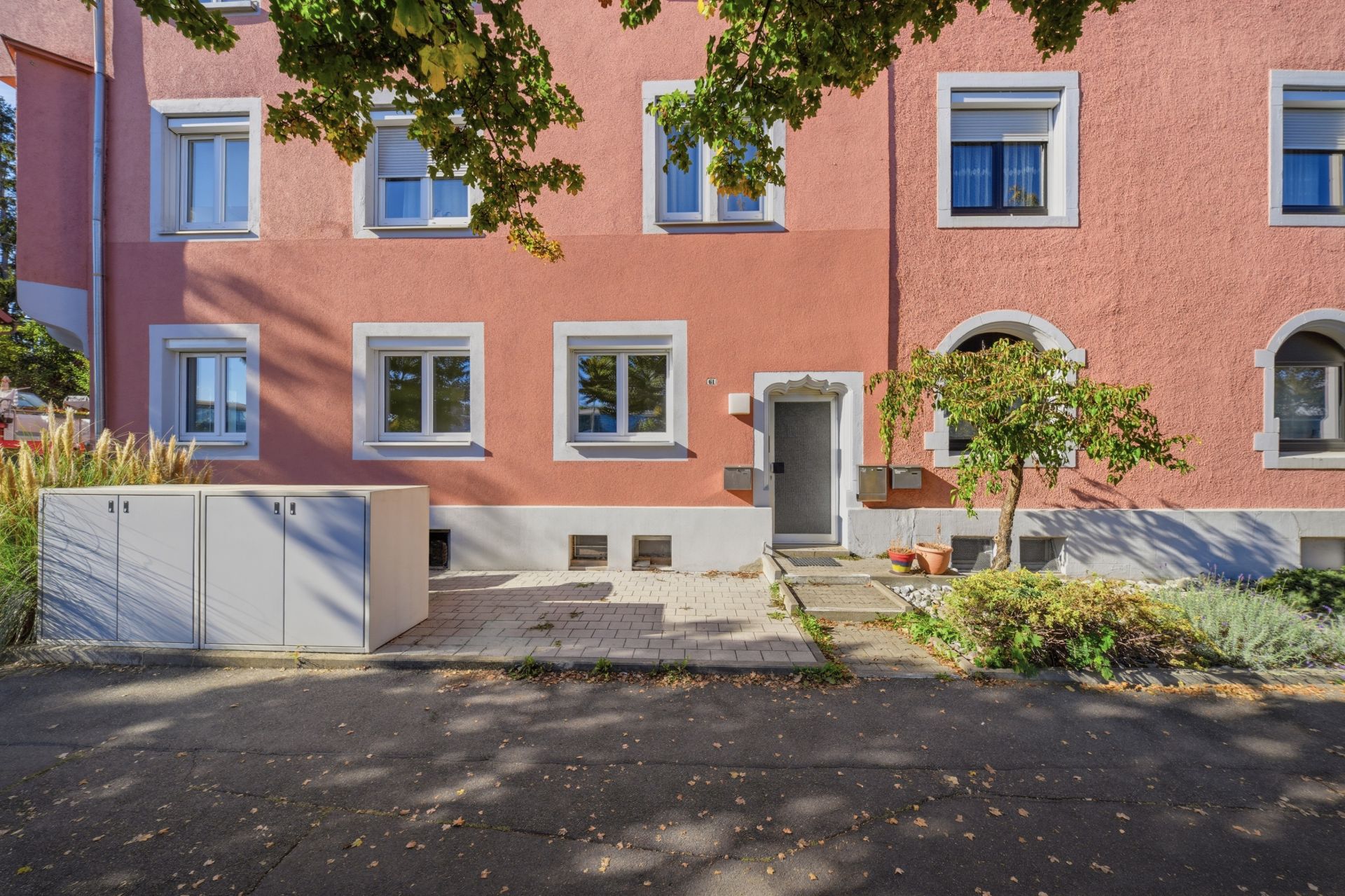Vorderansicht eines rosafarbenen Wohnhauses mit mehreren Fenstern und einem Baum vor dem Eingang.
