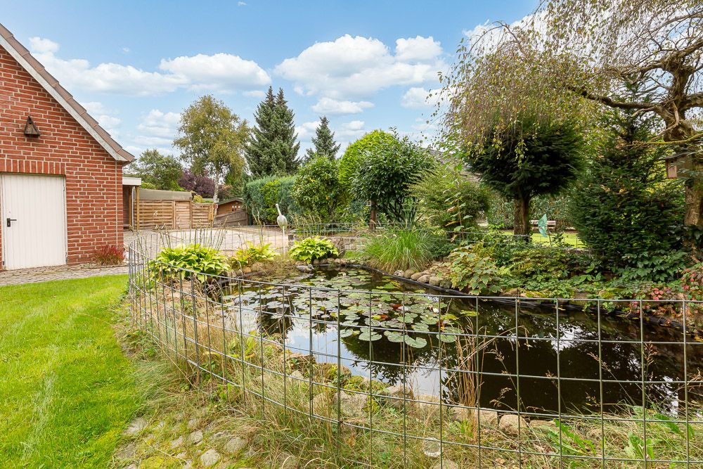 Garten mit kleinem Teich, Seerosen und Drahtzaun, umgeben von Bäumen und Sträuchern neben einem Backsteingebäude.