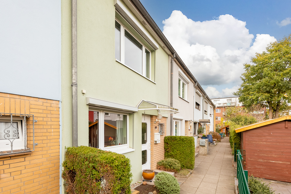 Reihenhaus mit grünem Busch vor dem Eingang und Gehweg entlang der Häuserreihe.