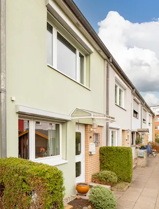 Reihenhaus mit grünem Busch vor dem Eingang und Gehweg entlang der Häuserreihe.