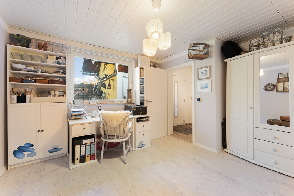 Helles Arbeitszimmer mit Schreibtisch, Regalen, Stuhl und großem Fenster, weiße Möbel und Deko.