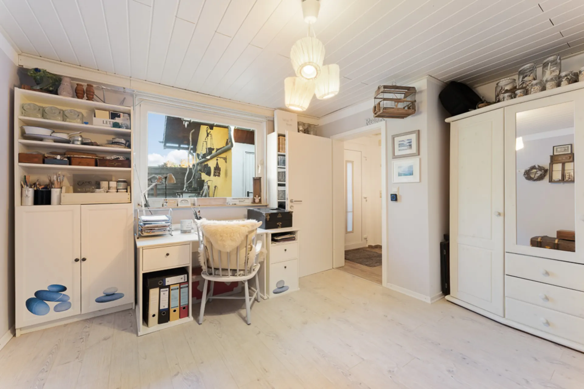 Helles Arbeitszimmer mit Schreibtisch, Regalen, Stuhl und großem Fenster, weiße Möbel und Deko.