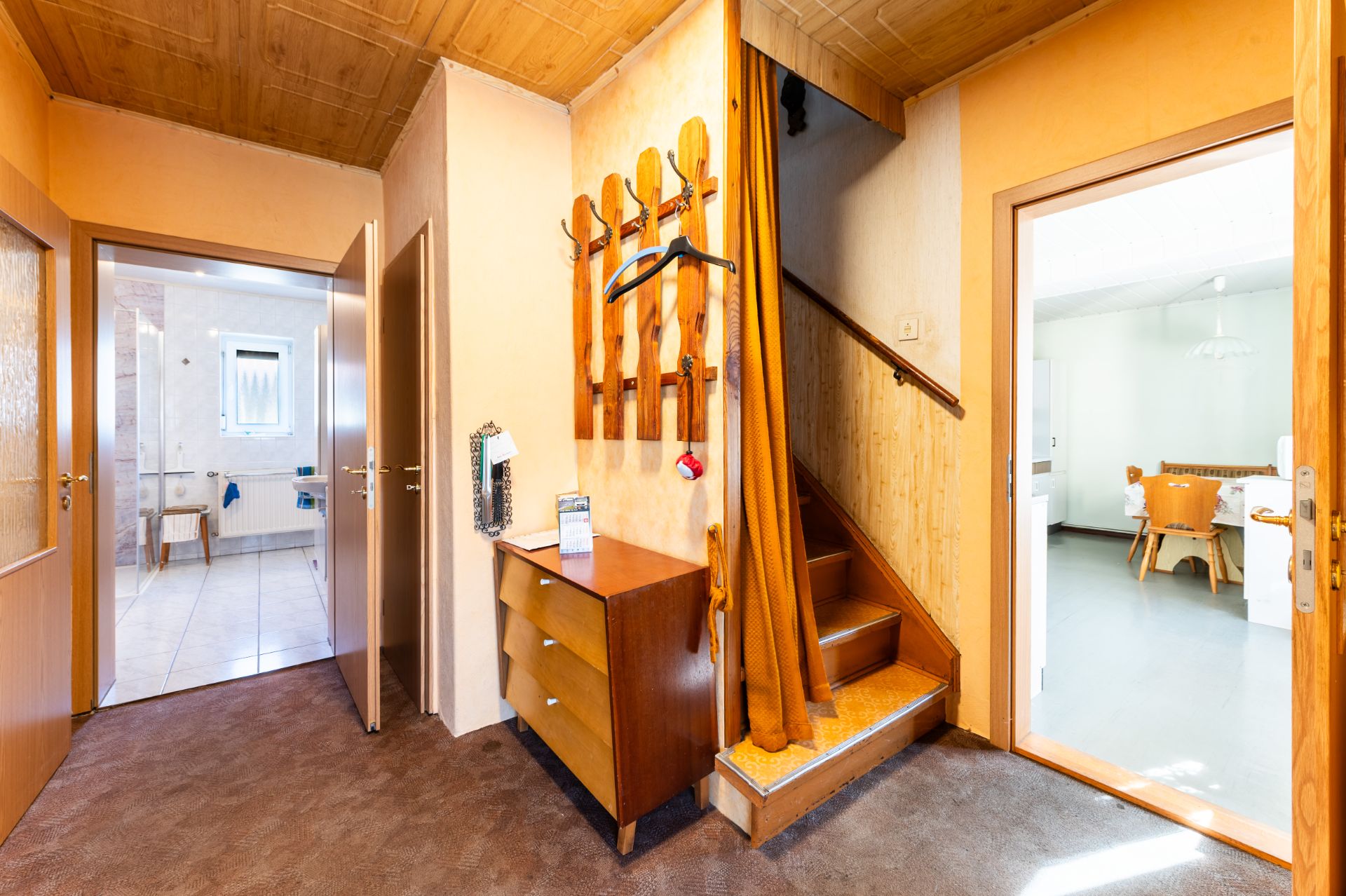 Flur mit Holzgarderobe, Kommode, Teppichboden, sichtbarer Treppe und Türen zu Bad und Esszimmer.