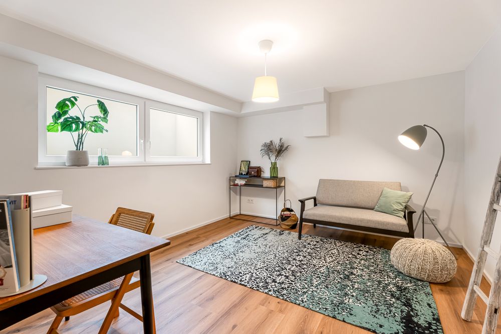 Helles Zimmer mit Schreibtisch, Sofa, Teppich und großen Fenstern, Holzboden und Zimmerpflanze.