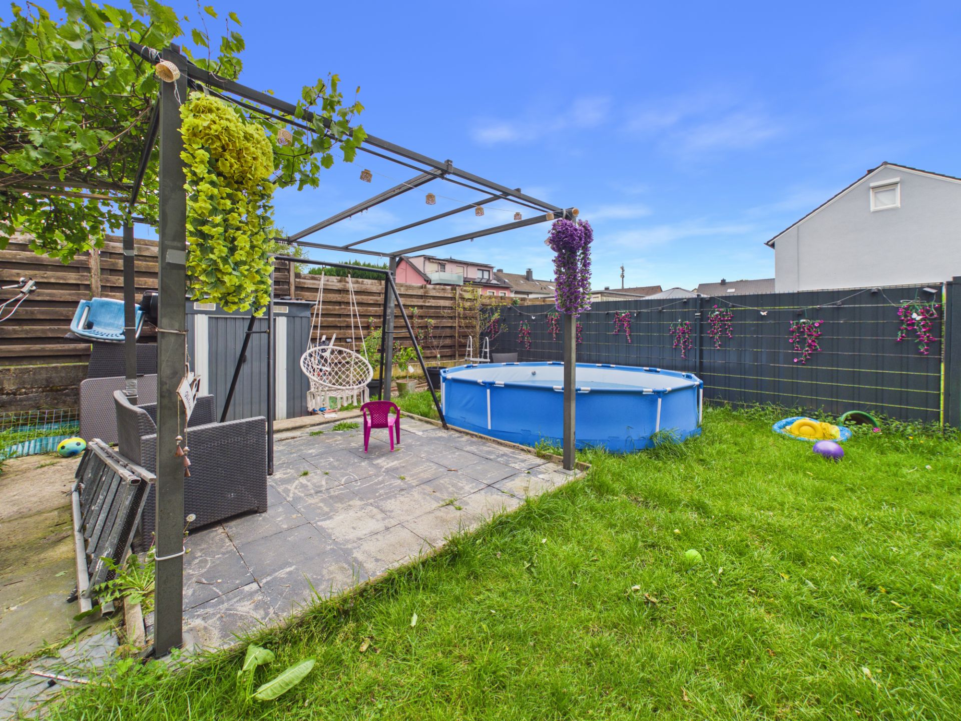 Garten mit Terrasse, Pergola, Sitzgruppe, Hängesessel, Planschbecken und Spielsachen auf dem Rasen.