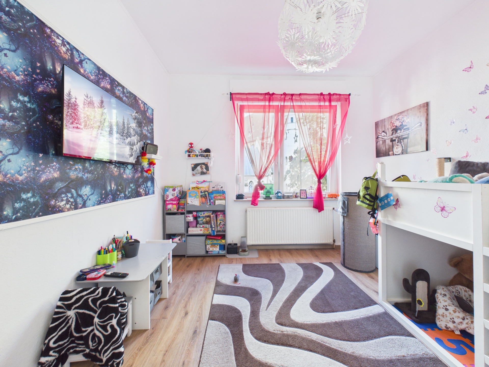 Helles Kinderzimmer mit Hochbett, Schreibtisch, Bücherregalen und bunten Vorhängen am Fenster.