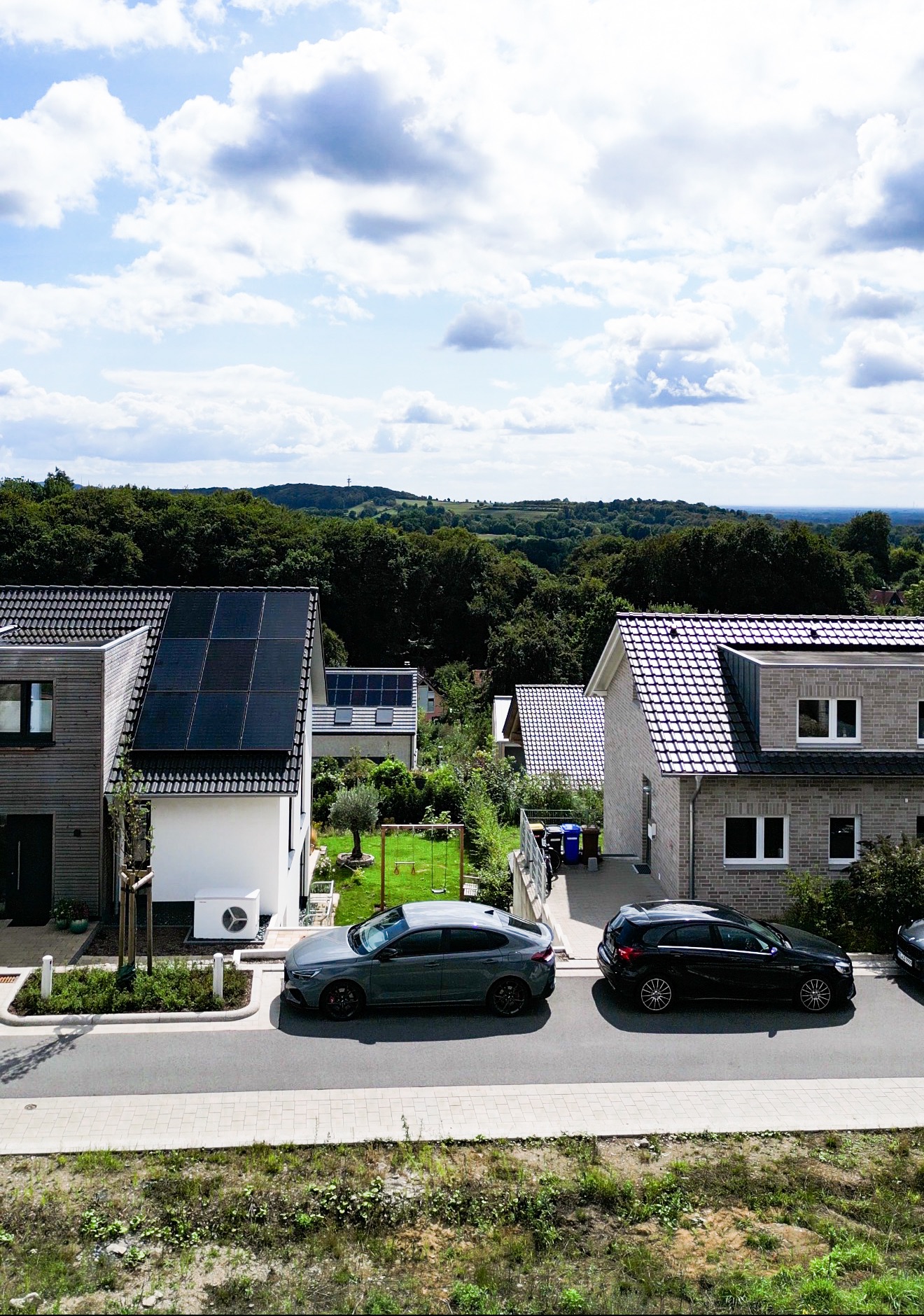 Zwei Einfamilienhäuser mit Solarpanels, Autos parken davor, im Hintergrund Wälder und Hügel.