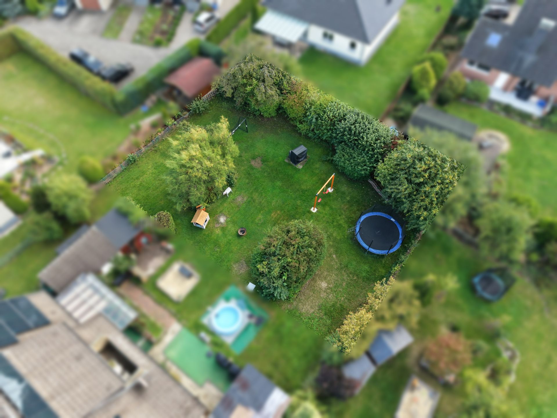 Luftaufnahme eines Gartens mit Trampolin, Bäumen, Spielhaus und Schaukel inmitten von Hecken.