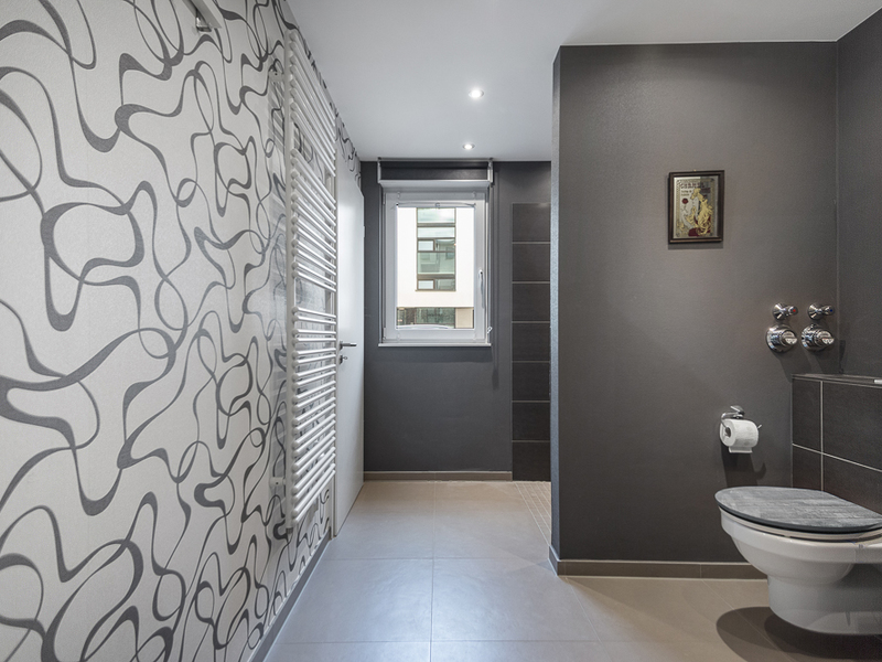 Modernes Badezimmer mit Wandtoilette, grauen Wänden, großem Fenster und gemusterter Tapete.
