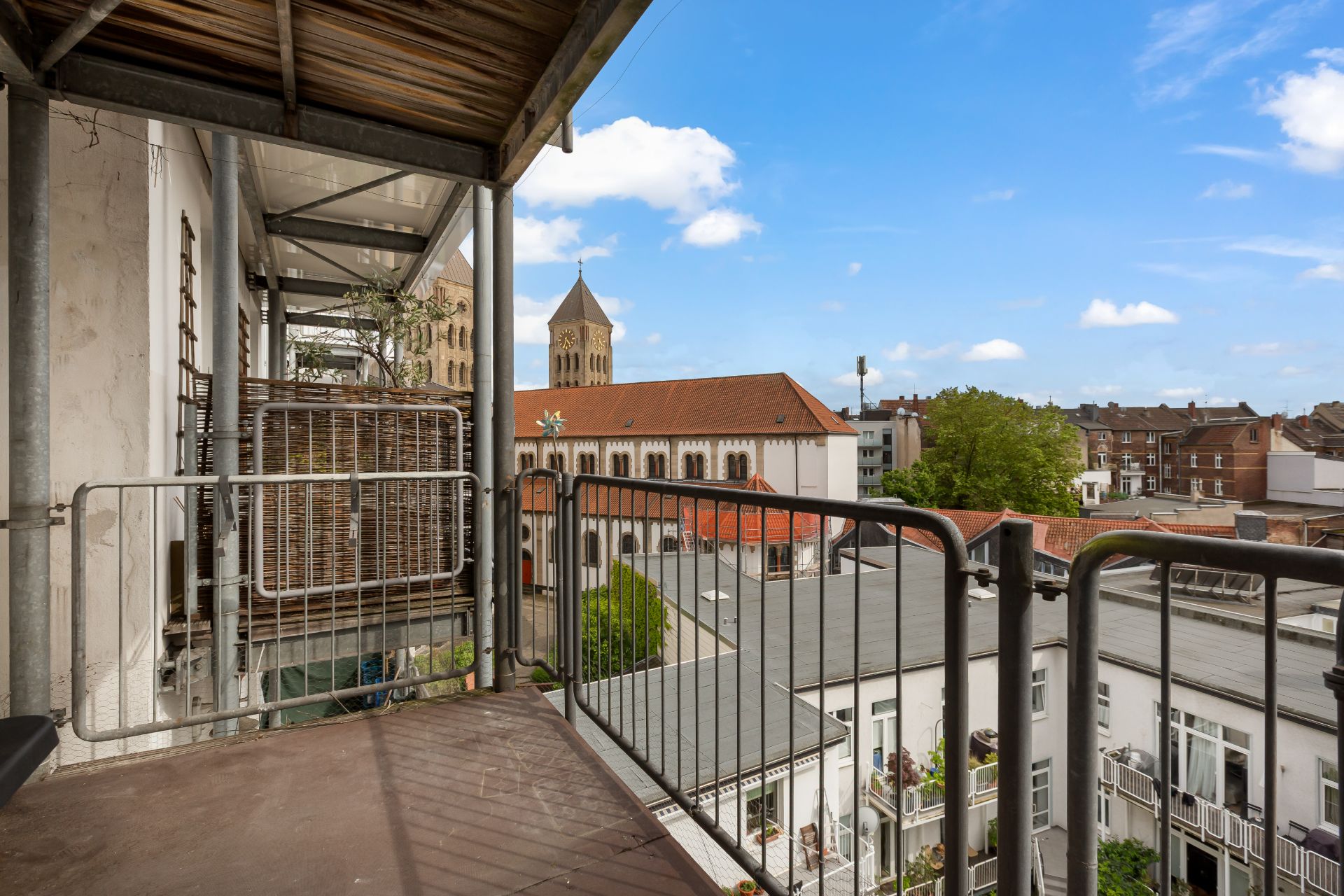 Balkon mit Metallgeländer, Blick auf umliegende Gebäude und eine Kirche im Hintergrund.