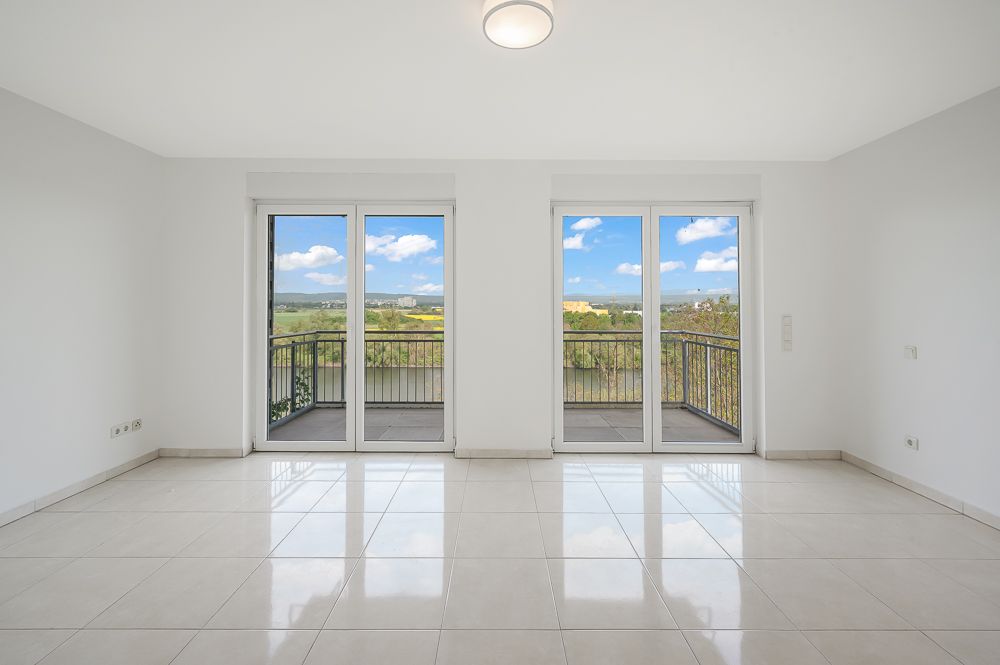 Helles Wohnzimmer mit Fliesenboden und zwei großen Glas­türen zu einem Balkon mit Ausblick.