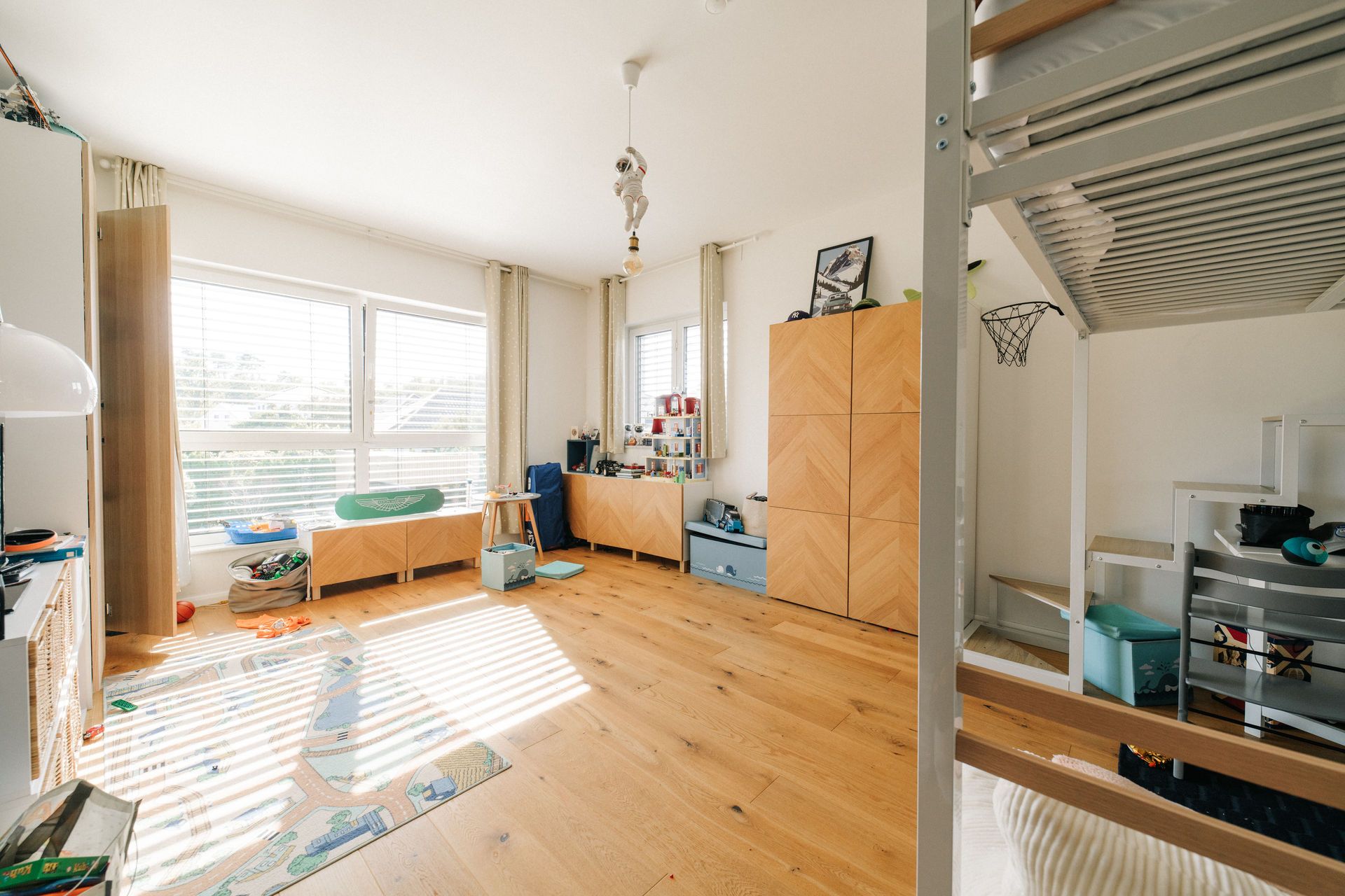 Helles Kinderzimmer mit Holzboden, Spielteppich, Hochbett und großen Fenstern.