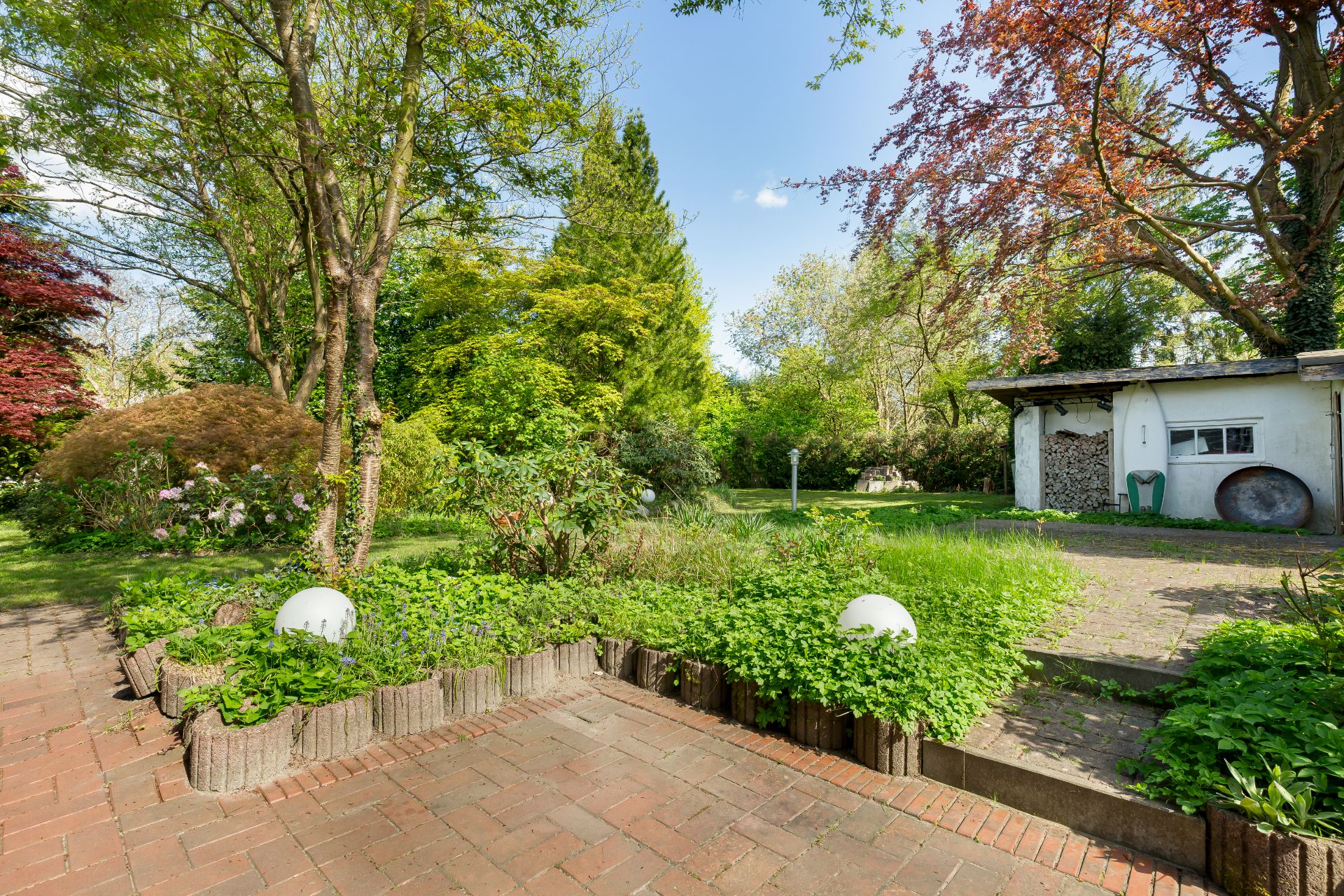 Gepflasterte Terrasse mit Garten, Bäumen, Rasenfläche und kleinem weißen Gartengebäude.