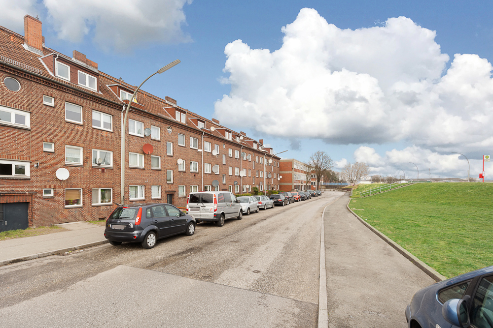 Rote Backsteinhausreihe mit geparkten Autos an einer ruhigen Straße und einer Wiese.