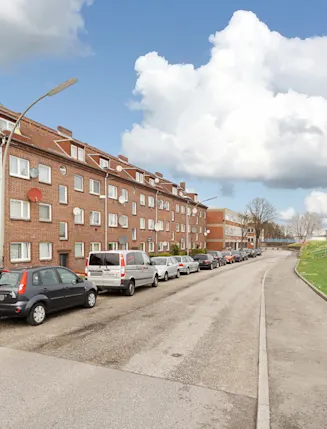 Rote Backsteinhausreihe mit geparkten Autos an einer ruhigen Straße und einer Wiese.