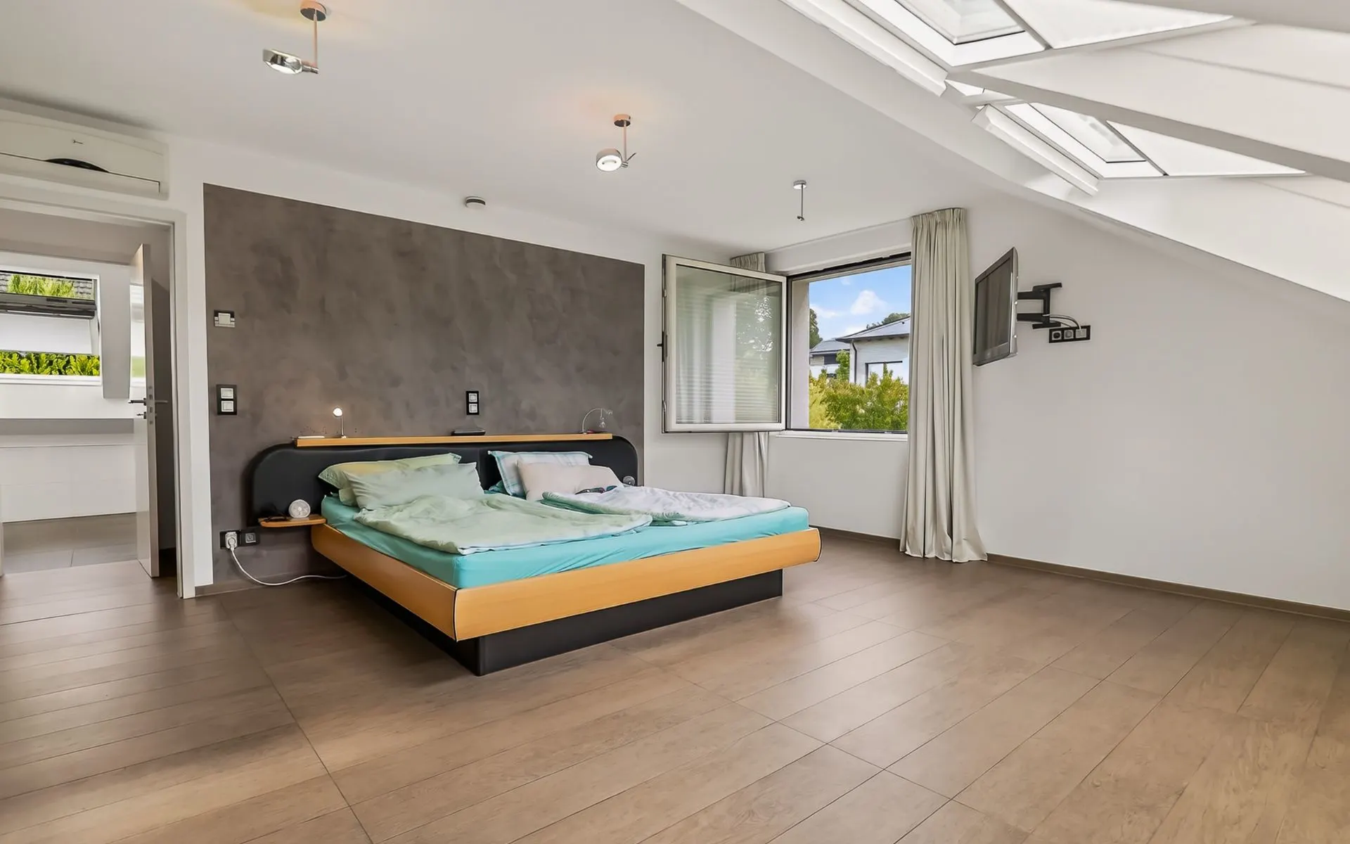 Helles Schlafzimmer mit Doppelbett, Holzboden, offenem Fenster und schräger Decke mit Dachfenstern.