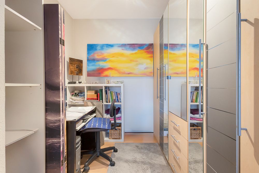Kleines Arbeitszimmer mit Schreibtisch, Drehstuhl, Regalen und buntem Wandbild.