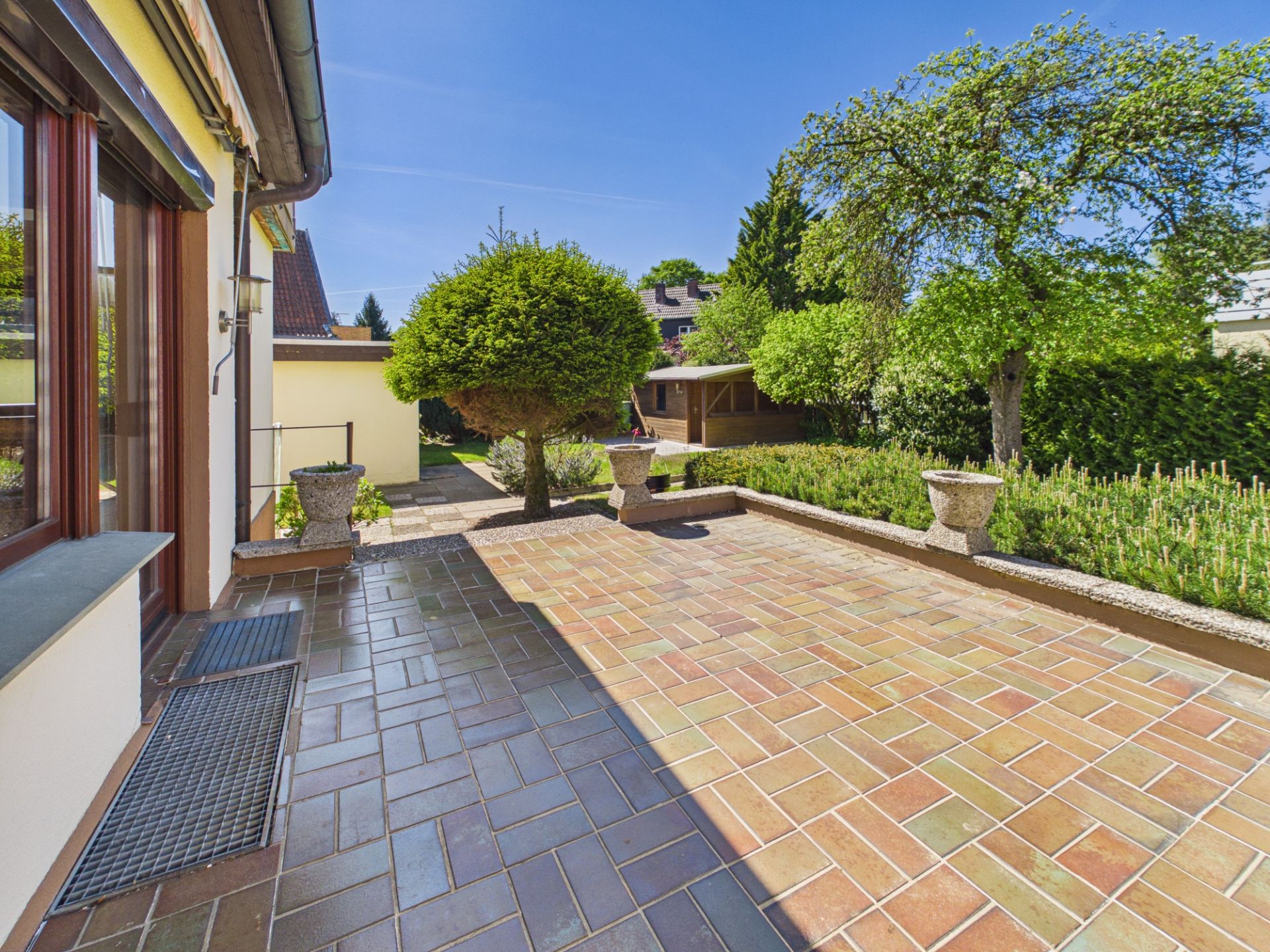 Große Terrasse mit Fliesenboden und Blick auf einen grünen Garten mit Bäumen und Blumenkübeln.