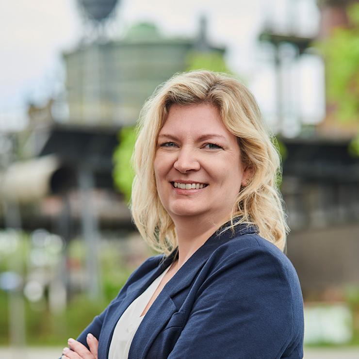 Frau mit blondem Haar und blauem Blazer steht draußen und lächelt in die Kamera.